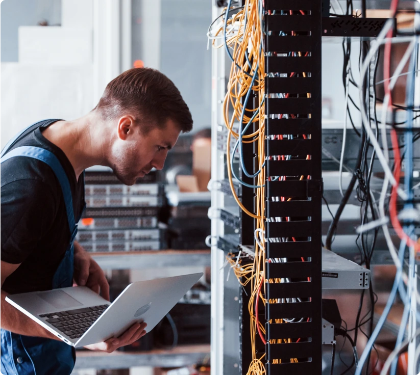 A man with a laptop in hand inspects a messy series of cables.