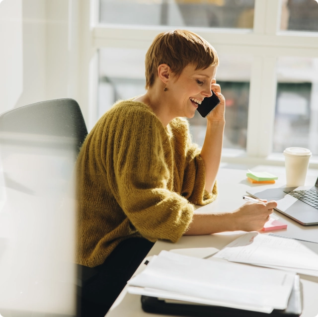 A woman writes on a sticky note and talks on the phone. She is laughing.