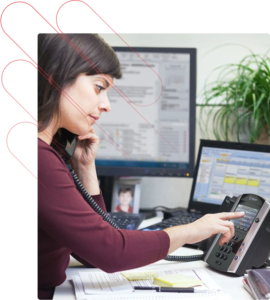 A woman dials on an office phone. There is a PC and a laptop on her desk.