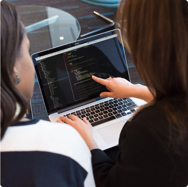 Two women sit in front of a laptop and look at code on the screen.